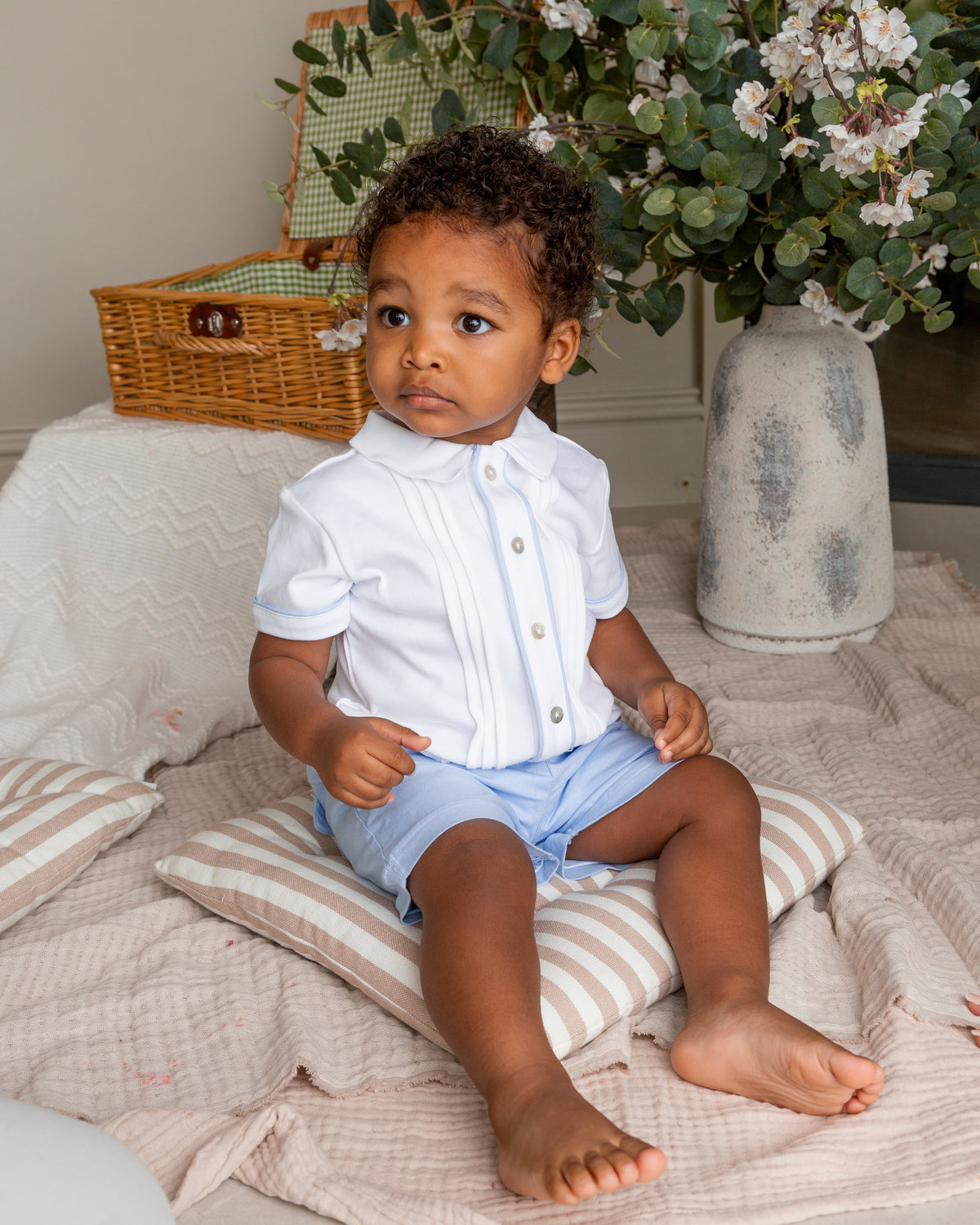 A toddler boy sitting on a striped cushion on a pink blanket, wearing a white short-sleeve button-up shirt with pleats and light blue shorts. The background features a wicker basket, a large vase with white flowers, and a neutral wall.