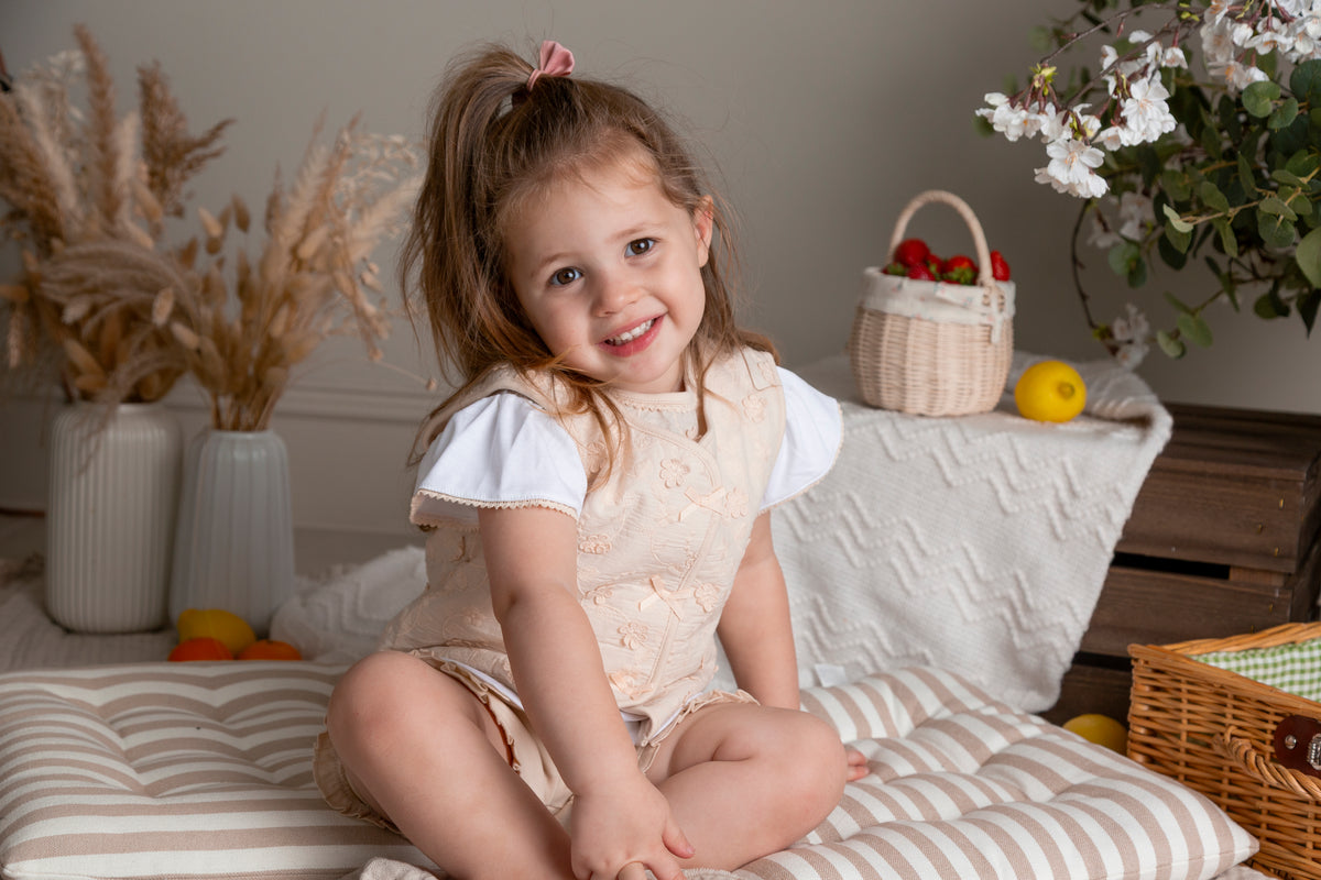 A young girl with a ponytail sits cross-legged on a striped cushion in a cozy indoor setting, wearing a cream-colored sleeveless dress over a white short-sleeve top. The background features dried pampas grass in vases, a basket of strawberries, and white flowers.